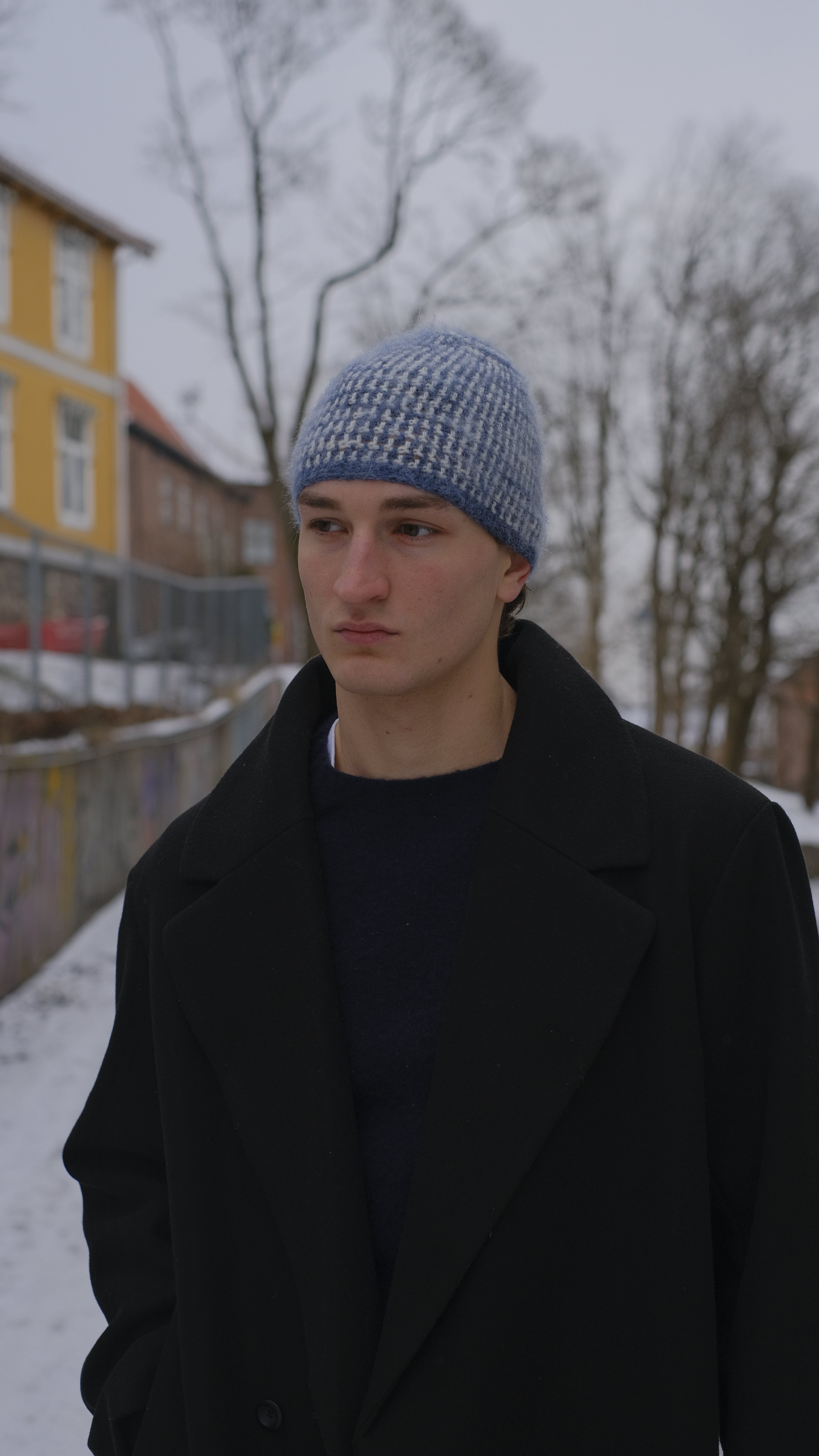 Person wearing a black coat and blue knit hat standing outdoors with snow on the ground.