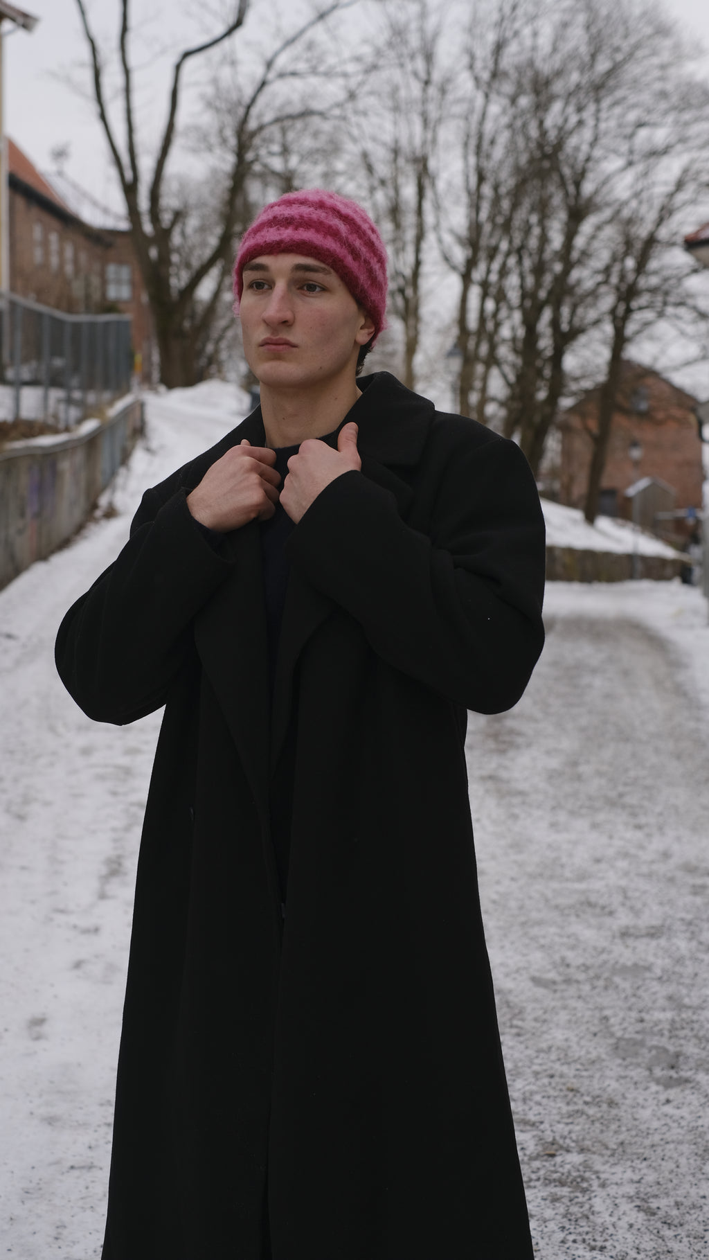 Person wearing a black coat and pink beanie standing in a snowy landscape with trees and buildings.