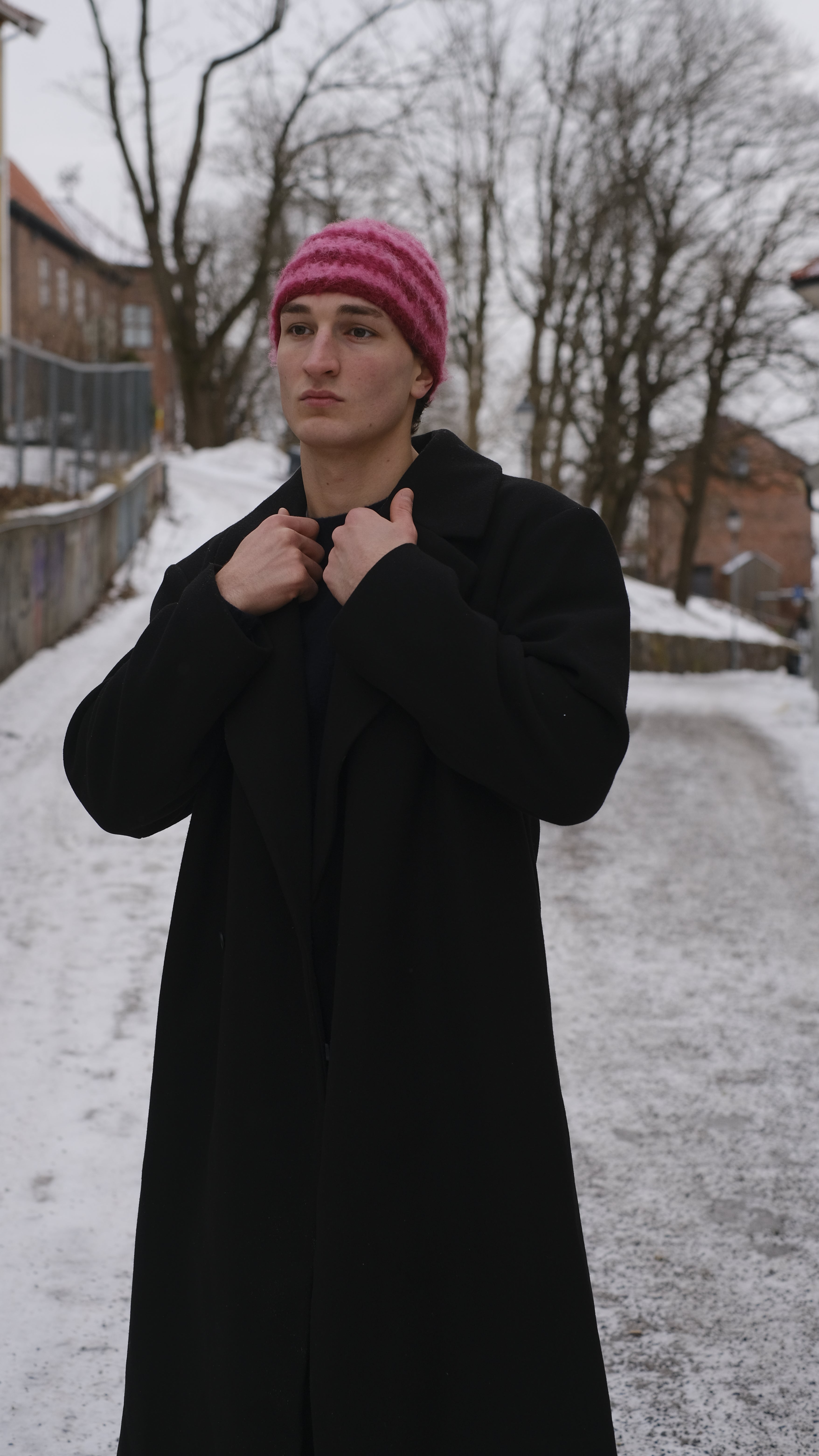 Person wearing a black coat and pink beanie standing in a snowy landscape with trees and buildings.