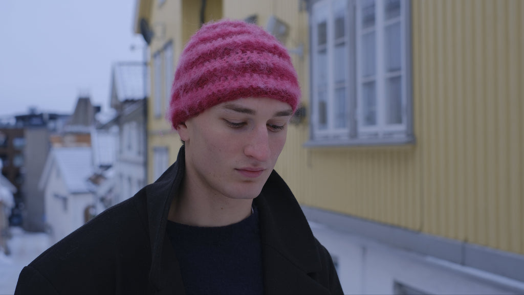 Person wearing a pink knit hat and dark coat standing in front of a yellow building with snow-covered roofs.